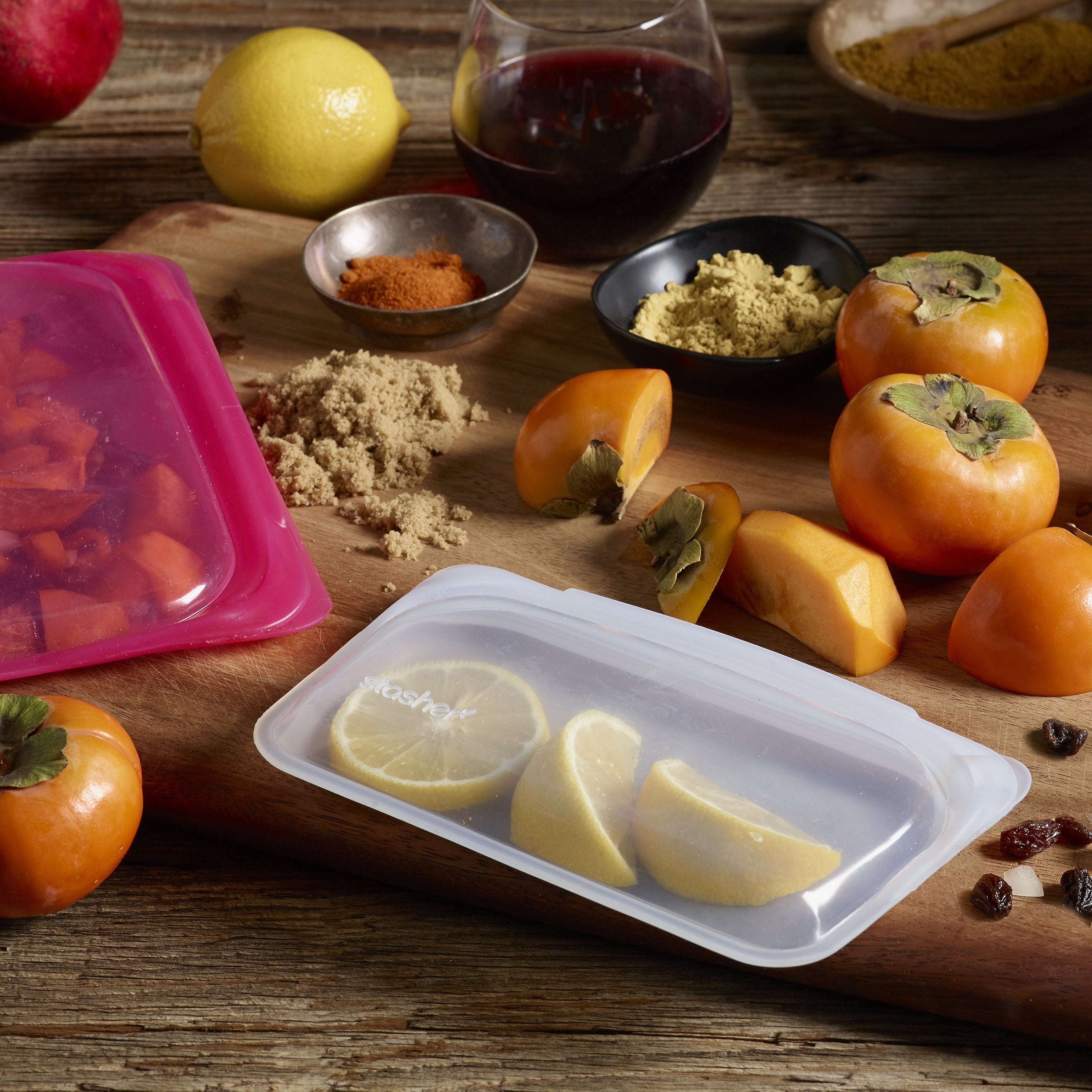 a clear snack sized stasher bag sitting on a wooden table that is holding lemon slices . There is also a pink stasher bag that is holding sliced tomatoes. There are fresh fruits, a glass of wine, and fresh spices sitting on the table next to the stasher bags.