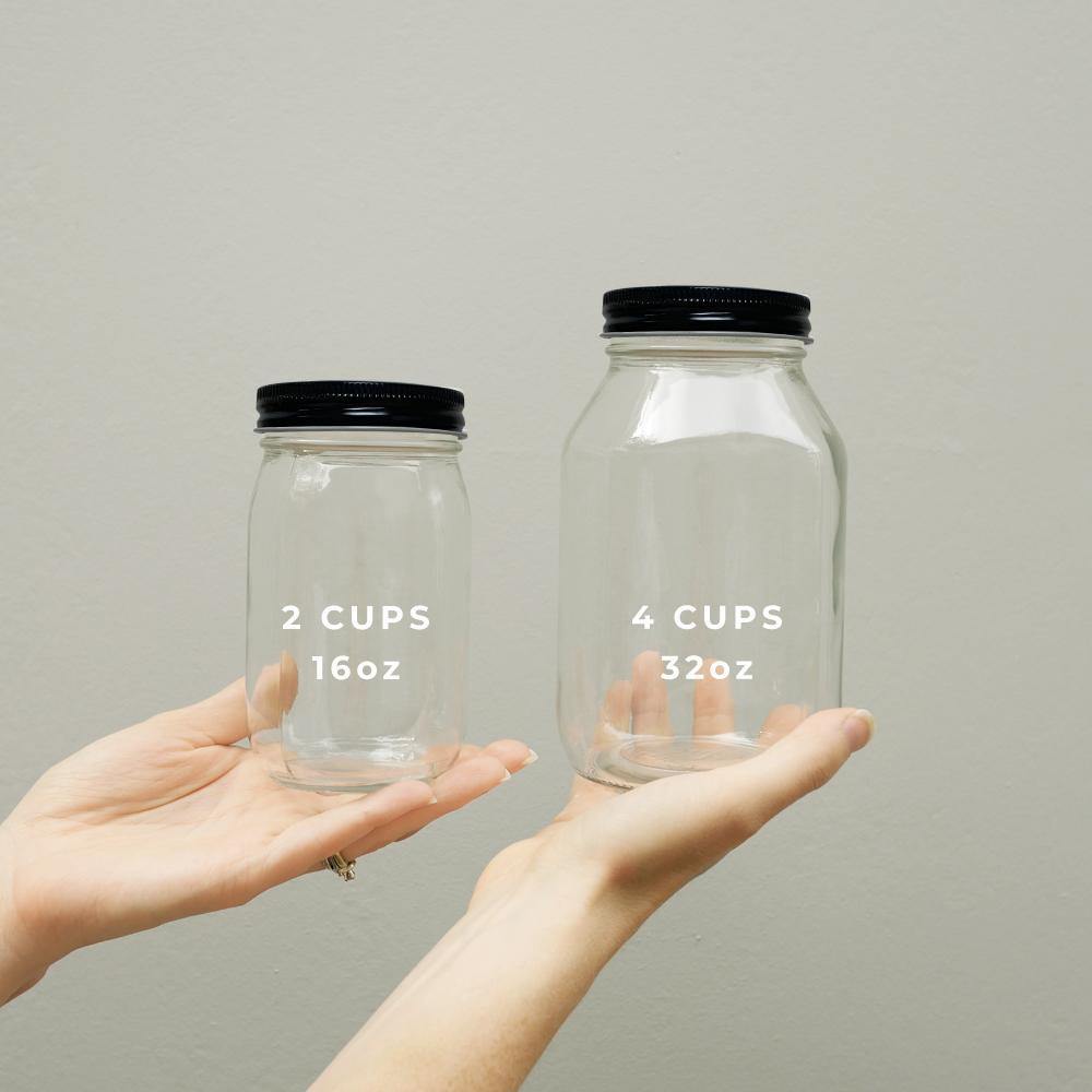 Hands holding two clear glass mason jars with black aluminum twist off lids. One jar is 16oz and one jar is 32oz.