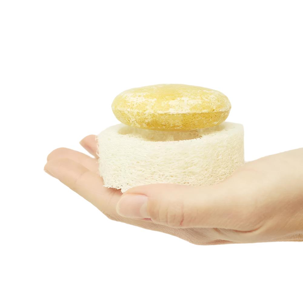 hand holding a round yellow shampoo bar that is sitting on top of a round zero waste loofah. The loofah is a natural white color.