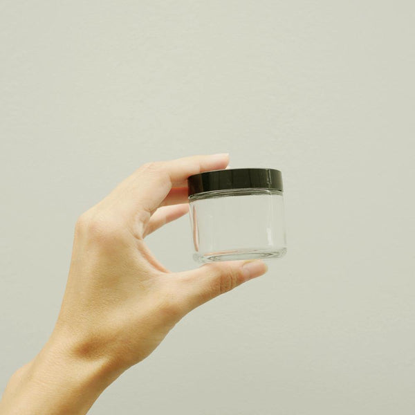 Hand holding an empty 2oz clear glass jar with a black twist off lid.
