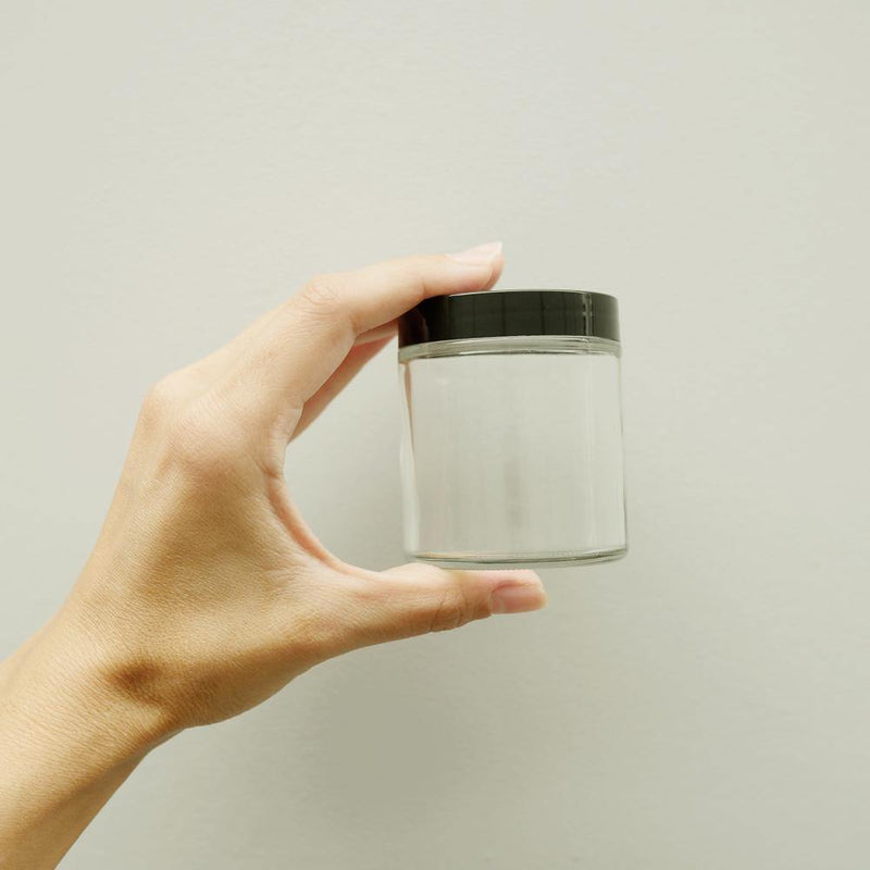 Hand holding an empty 4oz clear glass jar with a black twist off lid.
