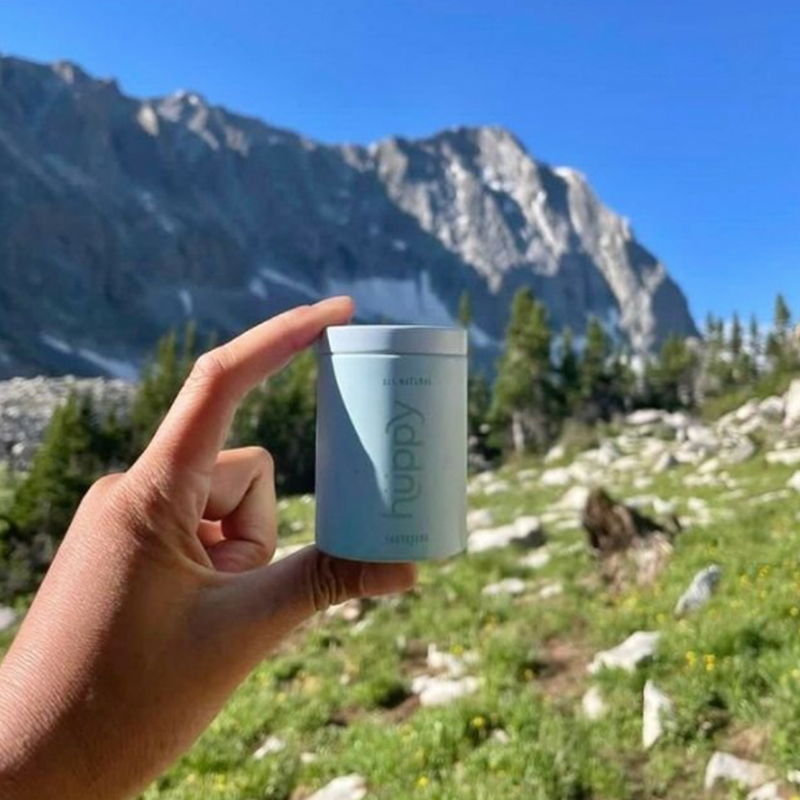 Someone holding a reusable huppy tin in front of a mountain scape.