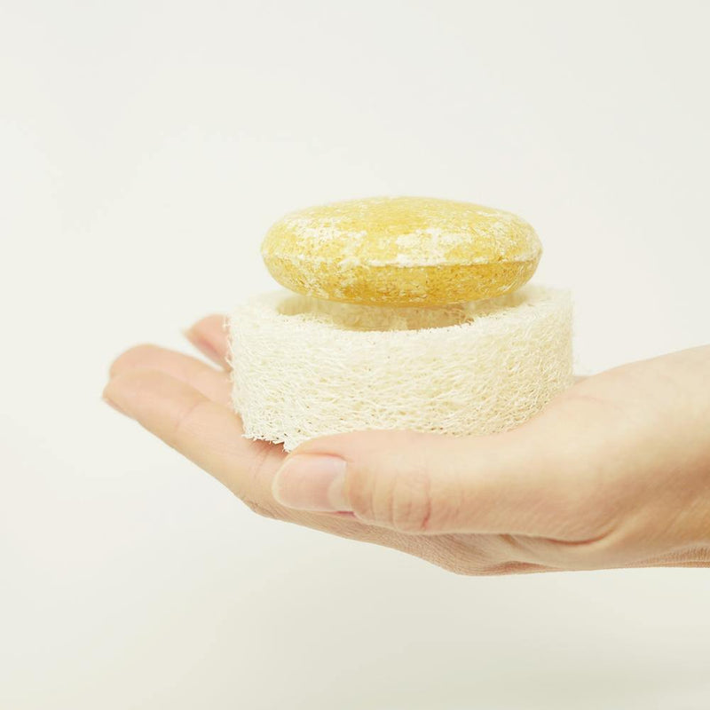 hand holding a round yellow shampoo bar that is sitting on top of a round zero waste loofah.  The loofah is a natural white color.