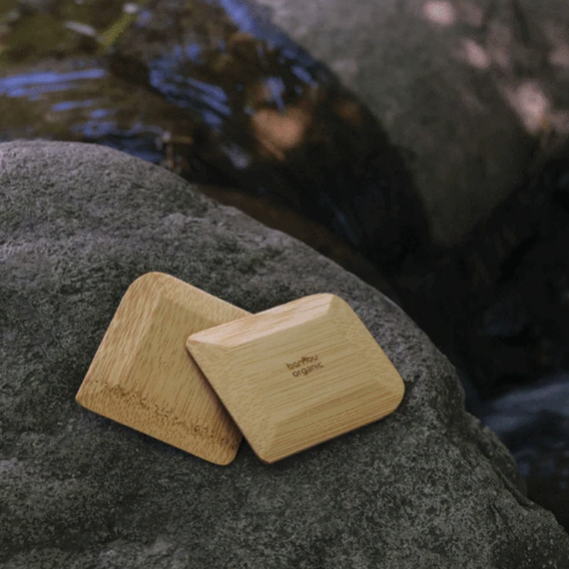 Two brown rectangular bamboo pot scrapers sitting on a rock beside a stream.
